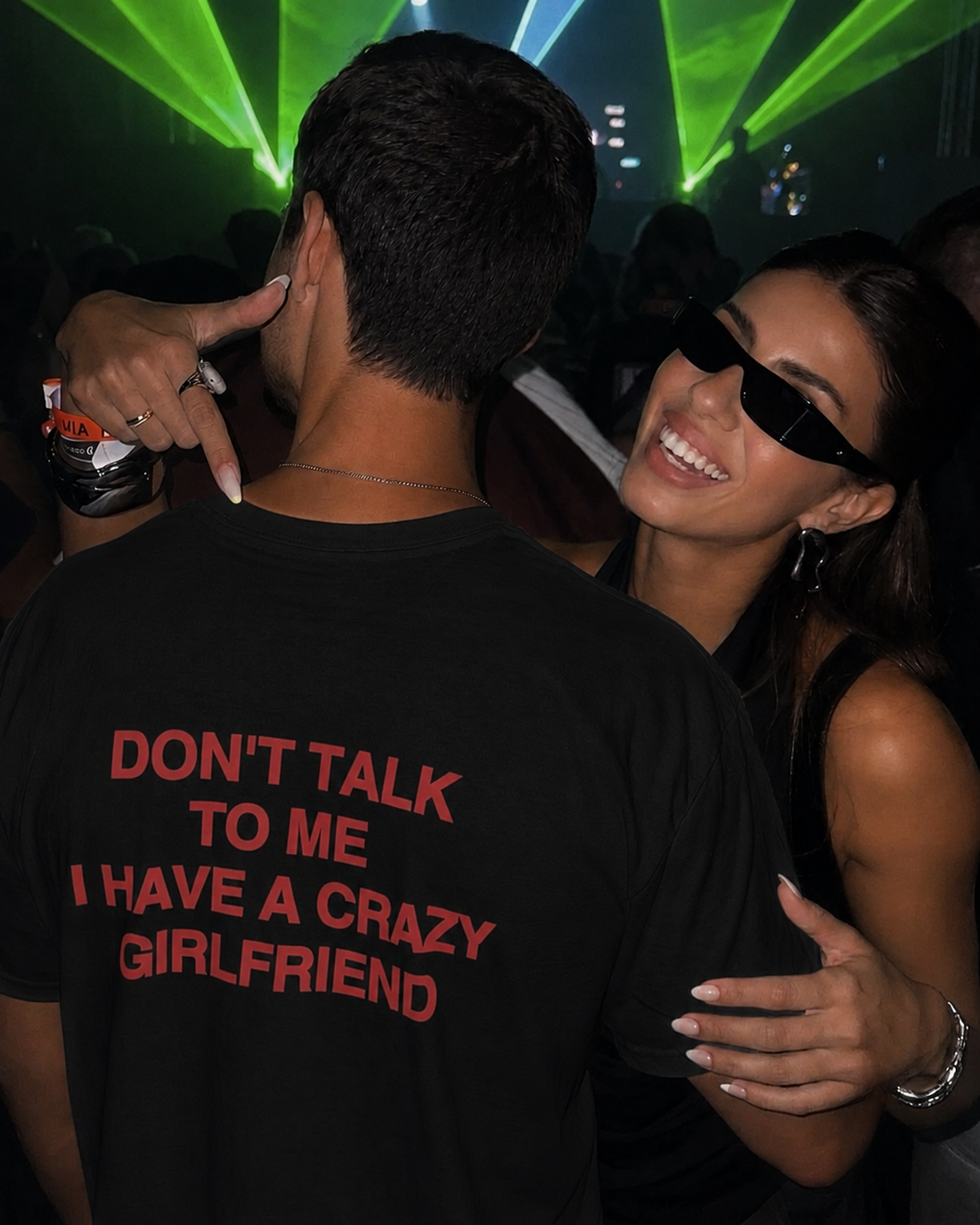 A man wearing a white graphic t-shirt with the text 'DON'T TALK TO ME I HAVE A CRAZY GIRLFRIEND' printed in red on the back, standing in a crowded nightclub with a woman wearing black sunglasses and a black top.