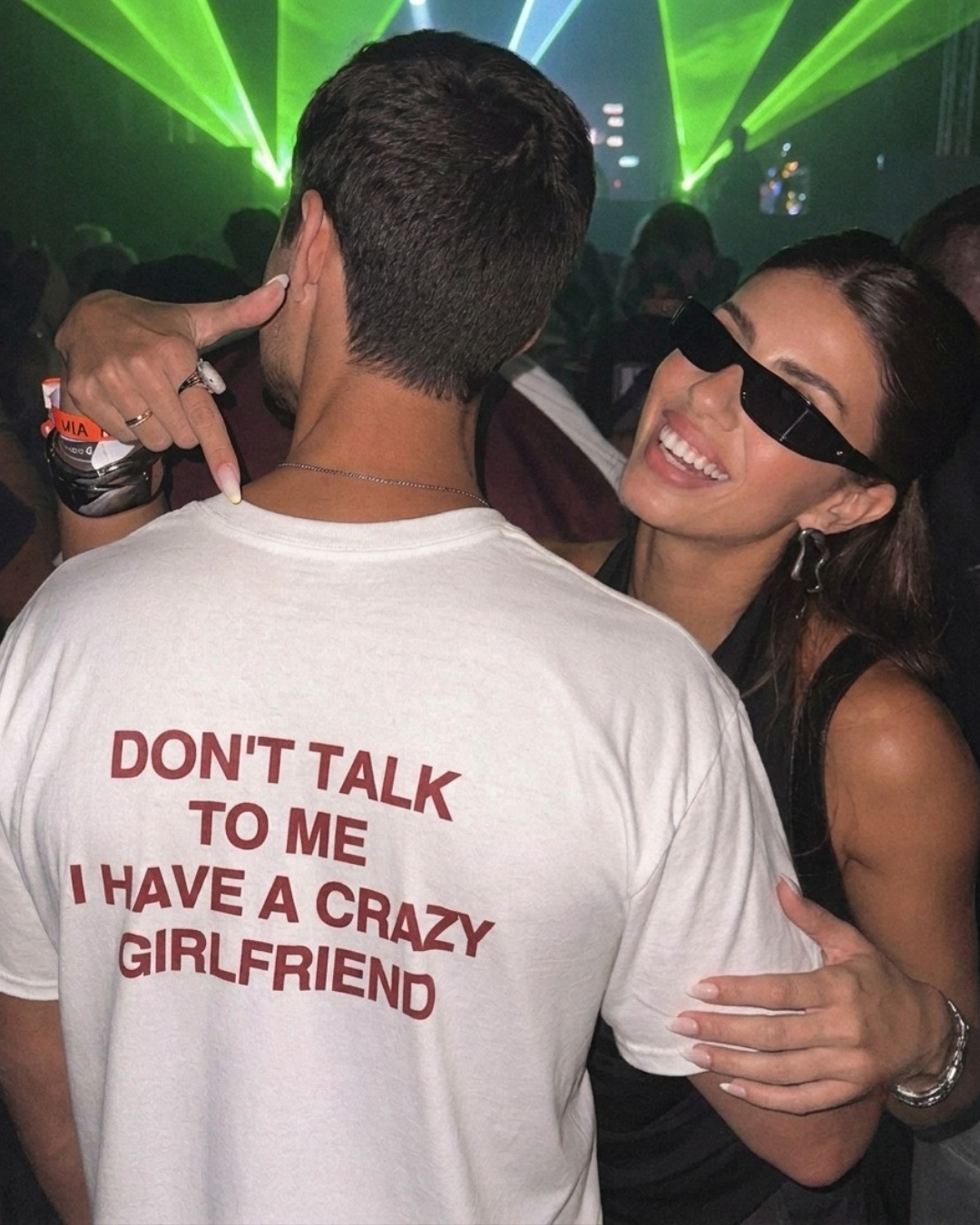 A man wearing a white graphic t-shirt with the text 'DON'T TALK TO ME I HAVE A CRAZY GIRLFRIEND' printed in red on the back, standing in a crowded nightclub with a woman wearing black sunglasses and a black top.